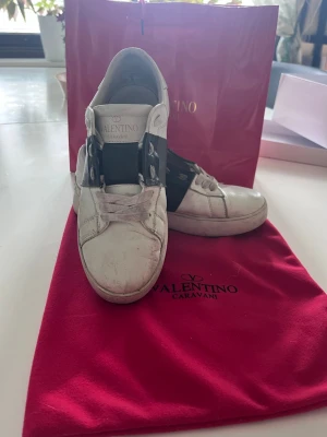 Valentino Garavani open vita sneakers - Snygga vita sneakers från Valentino Garavani med svart band över sidan och coola nitar. Tillverkade i läder och har en klassisk låg profil. Perfekt för dig som vill ha en exklusiv och trendig look.