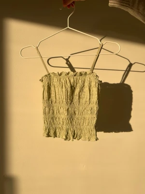 Ljusgrön smockad linnetopp - Trendig ljusgrön linnetopp med smockad struktur och tunna axelband. Toppen är croppad och har en mjuk, stretchig passform som sitter snyggt på kroppen. Perfekt för varma dagar och enkel att styla med jeans eller kjol.