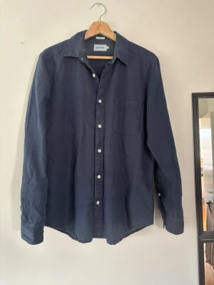 Mörkblå linneskjorta från East West - Snygg mörkblå skjorta från East West i linne. Skjortan har klassisk krage, vita knappar och en bröstficka. Perfekt för dig som gillar en enkel och stilren look. Långärmad och med en relaxed passform.