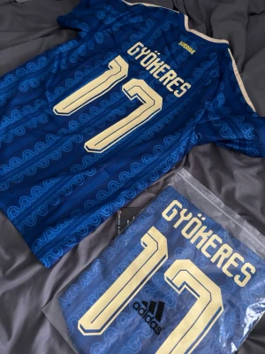 Blå Sverige fotbollströja Adidas - Säljer en blå Sverige fotbollströja från Adidas med Gyökeres och nummer 17 på ryggen. Tröjan har guldfärgade detaljer, korta ärmar och ett vågigt mönster över hela tyget. Perfekt för dig som vill stötta landslaget med stil.