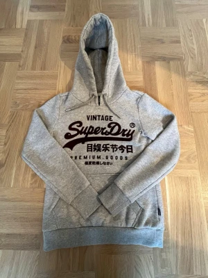 Grå superdry hoodie med tryck  - En riktigt fet grå superdry hoodie med ett snyggt tryck. Den är använd ett antal gånger men är i ett väldigt bra skick. Den är storlek Eu 36 enligt lappen med det motsvarar xs, något liten i storleken. Skriv gärna vid funderingar och priset är ej hugget i sten! 