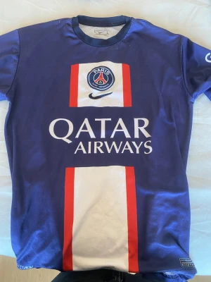 PSG matchtröja blå Nike - Säljer en blå Paris Saint-Germain matchtröja från Nike med vita och röda detaljer. Tröjan har PSG-logga på bröstet, Qatar Airways som sponsor och GOAT på ärmen. Materialet är lätt och andas, perfekt för fotboll eller träning.