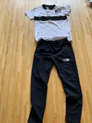 Svart och vit set från The North Face - Säljer ett snyggt set från The North Face med en vit t-shirt med svart band över bröstet och svarta byxor med logga. T-shirten har korta ärmar och knappar vid halsen, byxorna har resår i midjan. Perfekt för dig som gillar sportig stil.