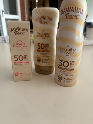 Hawaiian Tropic solskydd SPF 30/50 - Säljer tre solskyddsprodukter från Hawaiian Tropic: Glowing Protection Face Cream SPF 50, Hydrating Protection Lotion SPF 50 och Hydrating Protection Lotion Spray SPF 30. Produkterna är i beige och guld med vita detaljer och är avsedda för återfuktning och skydd mot solen.