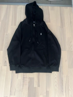 Svart hoodie från Polo Ralph Lauren - Svart hoodie med dragkedja från Polo Ralph Lauren. Klassisk design med huva och snören, två stora fickor framtill och den ikoniska vita Polo-loggan på bröstet. Tillverkad i mjukt bomullsmaterial som är perfekt för en avslappnad och stilren look.