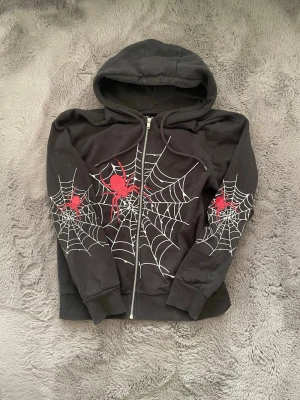Svart hoodie med spindelnätsmönster - Cool svart hoodie med dragkedja och huva. Framsidan och ärmarna har vita spindelnät och röda spindlar som ger en edgy vibe. Perfekt för dig som gillar unika och lite alternativa plagg. Muddar vid ärmslut och nederkant för extra komfort.