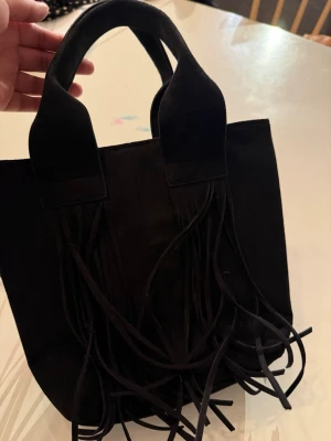 Svart handväska med fransar från Zara - Cool svart handväska från Zara med fransdetaljer på framsidan och stadiga handtag. Väskan är tillverkad i ett mjukt material som känns lyxigt och har en unik design med fransar som ger en trendig look. Perfekt för dig som vill sticka ut med din stil.