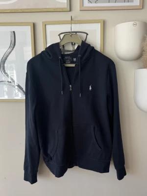 Marin blå Zip hoodie från Polo Ralph Lauren - Säljer en marin blå zip hoodie från Polo Ralph Lauren med dragkedja och klassisk logga broderad på bröstet. Hoodien har huva med snören, två stora fickor fram och ribbade muddar. Perfekt för en chill och stilren look. Pris diskuterbart 