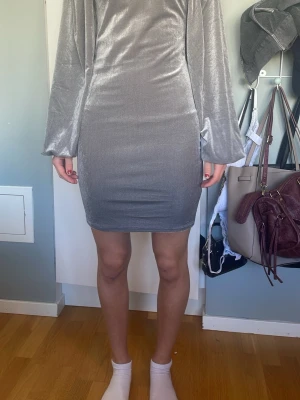 Grå kort klänning med ballongärmar - Säljer en snygg silver/grå kort klänning med ballongärmar. Klänningen har en tight passform och är gjord i ett glansigt, mjukt sammetmaterial som ger en lyxig känsla. Perfekt för dig som vill ha något stilrent och trendigt.