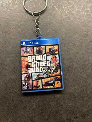 Nyckelring GTA V PS4-fodral - Säljer en unik nyckelring formad som ett PS4-fodral för spelet Grand Theft Auto V. Nyckelringen är i plast med detaljerad tryckt framsida och baksida, och har en metallkedja och ring. Perfekt för dig som gillar gaming och vill ha något personligt till nycklarna.