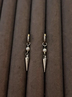 Silvriga örhängen med pärla och spike - Snygga örhängen i silverfärgad metall med en liten vit pärla och en spetsig spike-hängande detalj. Örhängena har en enkel ring som fäste och en cool, minimalistisk design som passar dig som gillar edgy accessoarer.