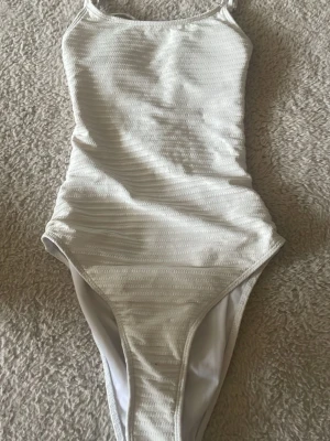 Vit baddräkt med fina detaljer  - Den är vit med mönster. Den har en fin öppen rygg och den knäppa med en silver knapp. Jätte fin när man är solbränd, poppar ut. 