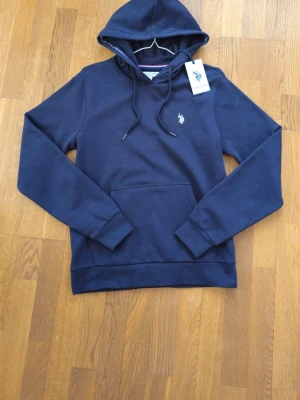 Mörkblå hoodie från U.S. Polo Assn. - Snygg mörkblå hoodie från U.S. Polo Assn. med klassisk huva och dragsnören. Tröjan har en broderad logotyp på bröstet och en stor magficka. Materialet är mjukt och bekvämt, perfekt för en avslappnad look. Helt ny oanvänd kan gå ner i pris vid snabb försäljning.