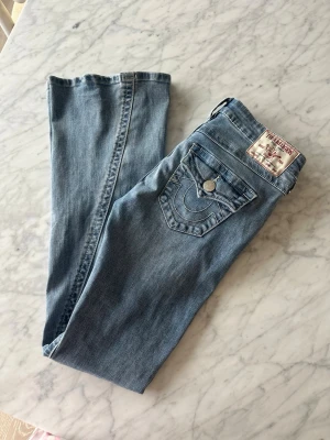 Ljusblå True Religion jeans - Säljer ett par ljusblå jeans från True Religion med ikonisk ficka och kontrastsömmar. Klassisk femficksmodell med knappdetalj och patch bak. Jeansen har rak passform och är tillverkade i mjuk denim för en avslappnad look.
