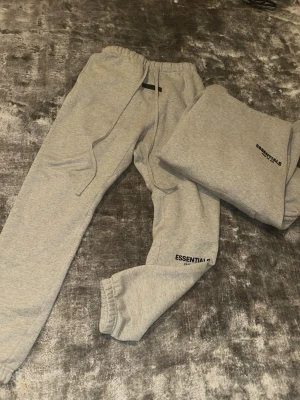 Essentials Tracksuit - Säljer ett par gråa mjukisbyxor från Fear of God Essentials. Byxorna har resår i midjan och benslut, samt dragsko för bästa passform. Tillverkade i mjukt bomullsmaterial för maximal komfort. Perfekta för en chill och avslappnad stil.