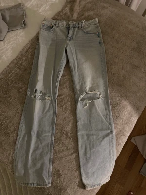 Ljusblåa straight fit jeans med slitningar - Säljer ett par ljusblåa jeans med straight fit och slitningar på båda knäna. Klassisk femficksmodell med knapp och dragkedja. Perfekt för dig som gillar en avslappnad och trendig look. Materialet är denim.