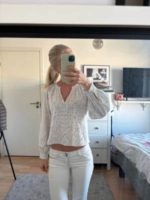 Vit broderad blus med lång ärm - Säljer en vit blus med broderade hålmönster och v-ringning. Blusen har en luftig passform och långa ärmar med resår vid ärmslut. Perfekt för dig som gillar romantiska detaljer och en fräsch look.