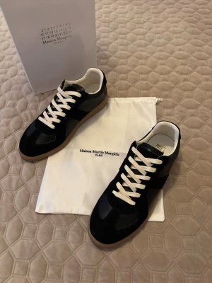 Maison margiela svarta sneakers - Maison Margiela sneakers i svart mocka och läder med vita skosnören och beige innersula. Klassisk låg modell med gummisula i brunt. Snygg och stilren design som passar till det mesta. Levereras med dustbag och originalkartong.