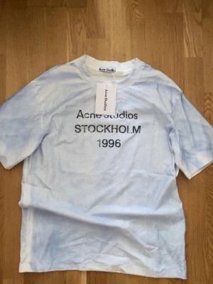 Acne Studios Stockholm T-shirt - Ljusblå och vit t-shirt från Acne Studios med trycket 'Acne Studios STOCKHOLM 1996' på bröstet. Oversized passform och korta ärmar. Tillverkad i mjuk bomull, perfekt för en avslappnad och trendig look.