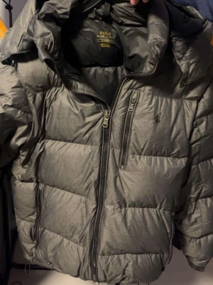 Grå dunjacka från Polo Ralph Lauren - säljer ralph lauren jacka då den inte längre kommer till användning. Storlek M. den har inga defekter eller tecken på användning. den är skrynklig eftersom den bara hängt vid massa jackor men det går bort vid användning!