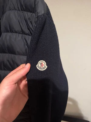 blå cardiganjacka från Moncler - Snygg blå cardiganjacka från Moncler med vadderad framsida och stickade ärmar. Jackan har dragkedja framtill, ståkrage och två fickor med dragkedja. Klassisk Moncler-logga på ärmen och ribbade muddar. Perfekt för lager-på-lager och streetstyle.