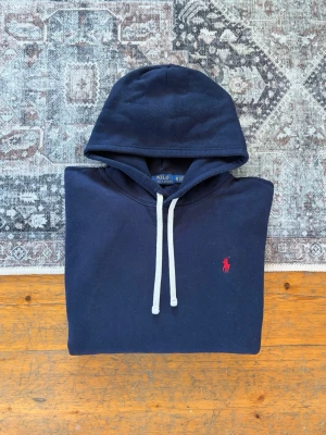 Marinblå Polo Ralph Lauren Fleece Hoodie - Storlek XL - Tjena! 👋 Säljer en klassisk marinblå (Navy) Polo Ralph Lauren hoodie i storlek XL. Den har den ikoniska röda pony-loggan broderad på bröstet – en tidlös färgkombination som alltid är rätt! 🐎 Tröjan är i mycket gott skick och välvårdad. Den är 100% äkta med både tvättlappar och QR-kod kvar för enkel verifiering ✅ Ett högkvalitativt basplagg som sitter perfekt och håller formen år efter år. Passar perfekt både för en rymligare passform eller om du har en större storlek. Fraktar snabbt! 🚛📦💬💯 