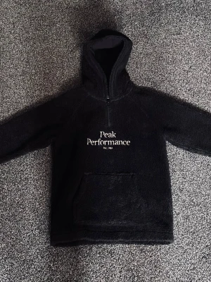 Svart fleecehoodie från Peak Performance - Mysig svart fleecehoodie från Peak Performance med stor logotyp på bröstet. Tröjan har halv dragkedja, stor huva och en praktisk magficka. Perfekt för kalla dagar och chill. Materialet är fluffigt och mjukt.