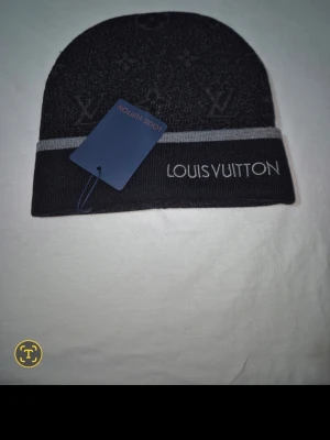 Svart mössa från Louis Vuitton - Snygg svart mössa från Louis Vuitton med diskret monogrammönster och grå kant. Märkets logga är broderad framtill. Tillverkad i mjukt material som känns lyxigt och bekvämt. Perfekt accessoar för dig som vill ha en exklusiv touch på din outfit.