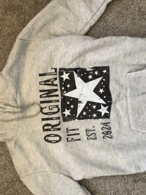Grå hoodie med stjärntryck - Säljer en ljusgrå hoodie från Original Fit med stor svartvit stjärna och texten 'ORIGINAL FIT EST. 2024' på framsidan. Hoodien har huva med dragsko och en klassisk magficka. Perfekt för dig som gillar streetstyle och coola prints. Om du brukar ha M så rekommenderar jag denna storlek eftersom denna L är en M egentligen (rep)