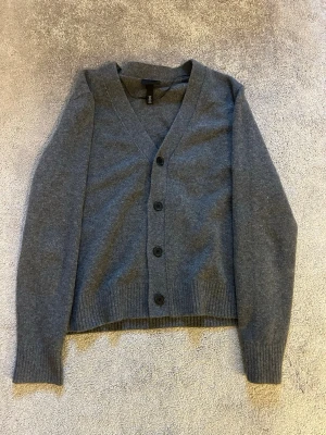 Mörkgrå stickad cardigan från H&M - Mysig mörkgrå stickad cardigan från H&M med v-ringning och fyra knappar framtill. Koftan har långa ärmar och ribbade muddar vid ärmslut och nederkant. Perfekt för lager på lager och enkel att matcha med olika outfits.