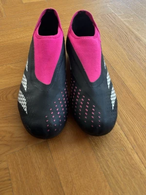 Svarta och rosa Adidas fotbollsskor - Säljer ett par snygga fotbollsskor från Adidas i svart och rosa med vita detaljer. Skorna har en strömlinjeformad design utan snörning och mönstrad ovandel för extra grepp. Perfekta för dig som vill sticka ut på planen med både stil och funktion.