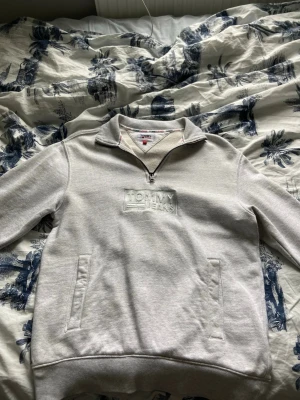 Ljusgrå quarterzip från Tommy Hilfiger - Snygg ljusgrå quarterzip hoodie från Tommy Jeans med broderad logga på bröstet. Tillverkad i 100% bomull och har två fickor framtill. Superbra skick och inga skador!