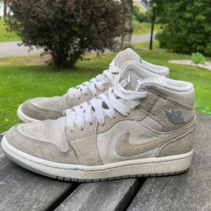 Nike Air Jordan 1 i beige mocka - Nike Air Jordan 1 sneakers i snygg beige och ljusgrå mocka med vita snören och klassisk Jordan-logga på sidan. Skorna har en rund tå, platt sula och ikonisk siluett som passar perfekt till streetwear. Mjukt material och stilren färg gör dem lätta att matcha.