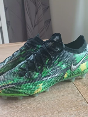 Nike Phantom GT2 Elite fotbollsskor - Säljer ett par Nike Phantom GT2 Elite fotbollsskor i grön, svart och gul färg. Skorna har en snygg mönstrad ovandel i syntetmaterial, snörning och dobbar för spel på konst gräs. Storlek 39.