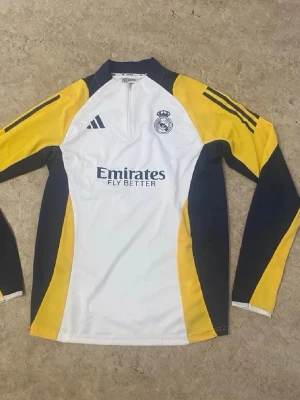 Real Madrid långärmad träningströja - Långärmad träningströja från Real Madrid med vit bas, gula och mörkblå detaljer. Tröjan har halv dragkedja framtill, klubbens logga på bröstet och Adidas-stripes på axlarna. Materialet är troligtvis polyester, perfekt för sport och träning.