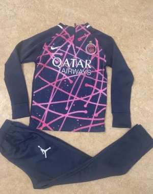 PSG träningsset från Jordan x Nike - Säljer ett PSG träningsset med långärmad tröja och matchande byxor. Tröjan är mörkblå med rosa och lila grafiskt mönster, PSG-logga och Qatar Airways-tryck. Byxorna är mörkblå med vit Jordan-logga. Perfekt för fotbollsträning eller chill.