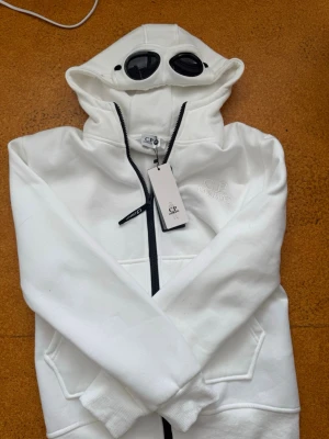 Vit hoodie med goggles från C.P. Company - Vit hoodie från C.P. Company med ikoniska goggles i huvan och svart dragkedja. Stora fickor framtill och broderad logga på bröstet. Perfekt för dig som vill sticka ut med en unik streetwear-look.