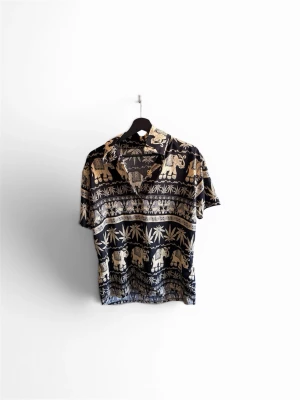 Svart skjorta med elefantmönster - Cool skjorta med korta ärmar och svart botten, dekorerad med beige elefanter och växtmönster. Skjortan har klassisk krage och knappar framtill, perfekt för dig som gillar unika prints och vill sticka ut. Köpt i Thailand!🙌🏼