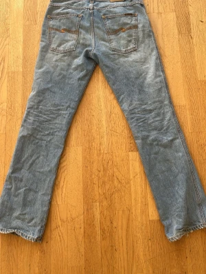 Blå Nudie Jeans Slim Jim  - Säljer ett par klassiska blå jeans från Nudie Jeans med bootcut och slitna detaljer vid benslut. Jeansen har fem fickor och snygga broderier på bakfickorna. Materialet är denim i bomull och passformen är rak med lätt utsvängda ben.