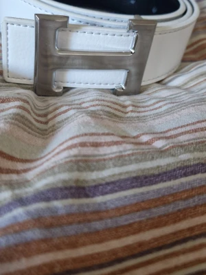 Vitt skärp med stor H-spänne - Snyggt vitt skärp med ett stort, blankt H-format metallspänne. Skärpet har stilrena sömmar och är gjort i ett slätt material som ger en clean look. Perfekt accessoar för att lyfta din outfit med en modern touch.