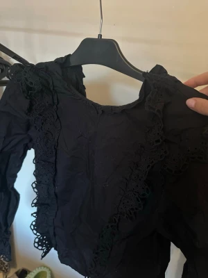 Svart blus med spetsdetaljer - Snygg svart blus med långa ärmar och dekorativa spetsdetaljer längs framsidan och axlarna. Blusen har knäppning framtill och en lätt rynkad struktur som ger en cool vintagekänsla. Perfekt för dig som gillar unika plagg med extra detaljer.