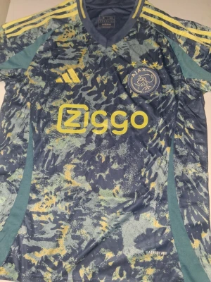 Ajax matchtröja camo Adidas - Ajax matchtröja från Adidas med camomönster i blått, grönt och gult. Tröjan har gula detaljer, Ajax klubbmärke på bröstet och Ziggo som sponsortryck. Tillverkad i lätt funktionsmaterial med AEROREADY-teknologi och klassisk sportig passform.