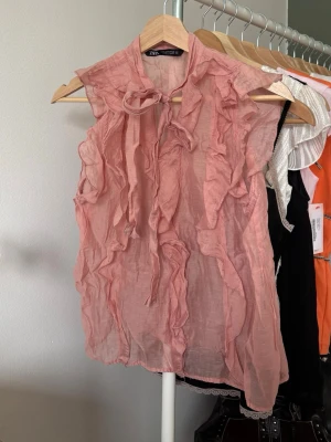 Rosa volangblus från Zara - Säljer en superfin rosa blus från Zara med volangdetaljer framtill och knytband vid halsen. Blusen är ärmlös och gjord i ett tunt material