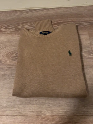 Beige stickad tröja Polo Ralph Lauren - Klassisk beige stickad tröja från Polo Ralph Lauren med rund halsringning och ribbade muddar. Tröjan har den ikoniska gröna broderade loggan på bröstet och är tillverkad i mjukt material som passar perfekt till lager-på-lager-stil.