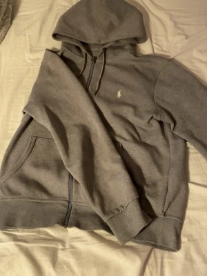 Polo zip hoodie - Klassisk grå hoodie från Polo Ralph Lauren med dragkedja, huva och fickor framtill. Ikoniska vita logon broderad på bröstet. Tillverkad i mjuk bomullsblandning som är perfekt för chill dagar eller när du vill ha en clean och enkel look. 