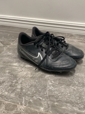 Nike Tiempo svarta fotbollsskor - Säljer ett par svarta Nike Tiempo fotbollsskor med klassisk vit swoosh på sidan. Skorna har snörning och fasta dobbar för spel på gräs. Ovandelen är i slitstarkt syntetmaterial och designen är stilren och sportig. Perfekta för dig som vill ha bra grepp och kontroll på planen.