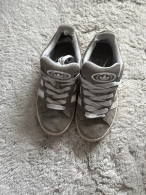Grå Adidas sneakers med vita detaljer - Säljer ett par gråa Adidas sneakers med klassiska vita ränder på sidorna och vit logga på plösen. Skorna har vita snören och en ljusbrun sula. Materialet är mocka och de har en cool retrostil som passar till många outfits.