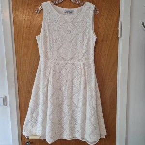 Vit spetsklänning med blommönster - Söt vit klänning i spets med blommigt mönster och ärmlös design. Klänningen har en rund halsringning och lätt utställd kjol som ger en feminin siluett. Perfekt för sommarens alla tillfällen. Off-white.