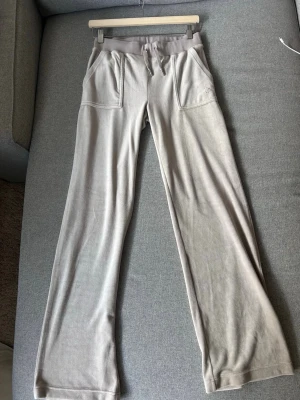 Beige mjukisbyxor Juicy Couture - Säljer ett par beige mjukisbyxor från Juicy Couture med raka ben, dragsko i midjan och fickor framtill. Byxorna har en liten Juicy Couture-logga broderad på ena fickan och är gjorda i ett mjukt, stretchigt material som är riktigt skönt.