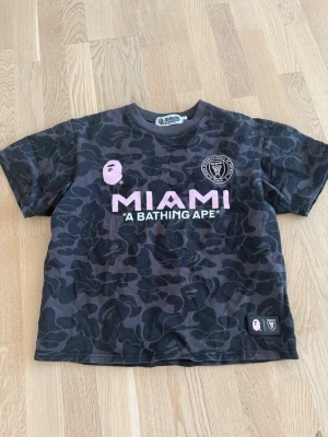 Svart camo BAPE x Miami T-shirt - Snygg svart camo T-shirt från BAPE x Miami med rosa tryck fram och bak. Stor MIAMI-logga och BAPE-ansikte på bröstet, samt stort 93-tryck och BAPE-text på ryggen. Kortärmad modell i mjuk bomull, perfekt för dig som gillar streetwear.